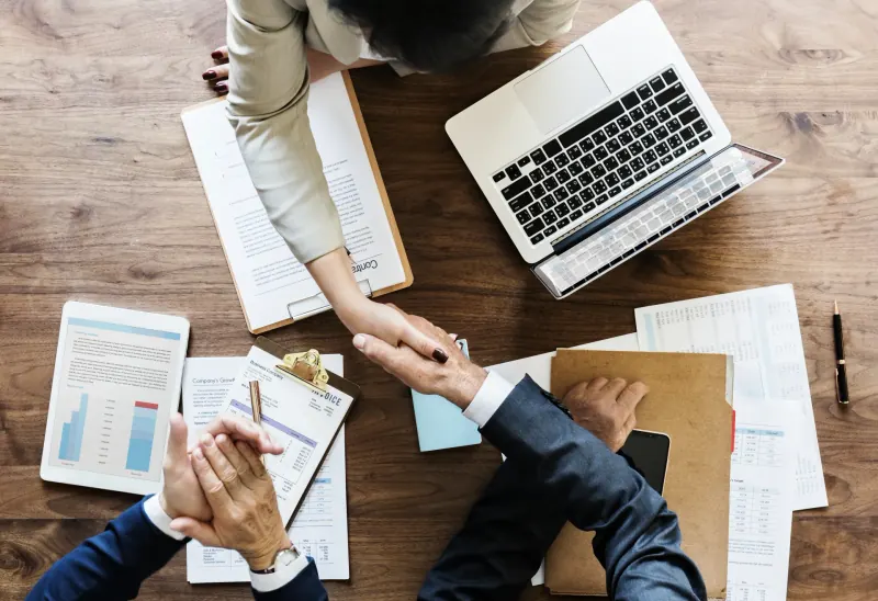 Overhead view of two people shaking hands across a desk covered in paperwork, a laptop and a tablet