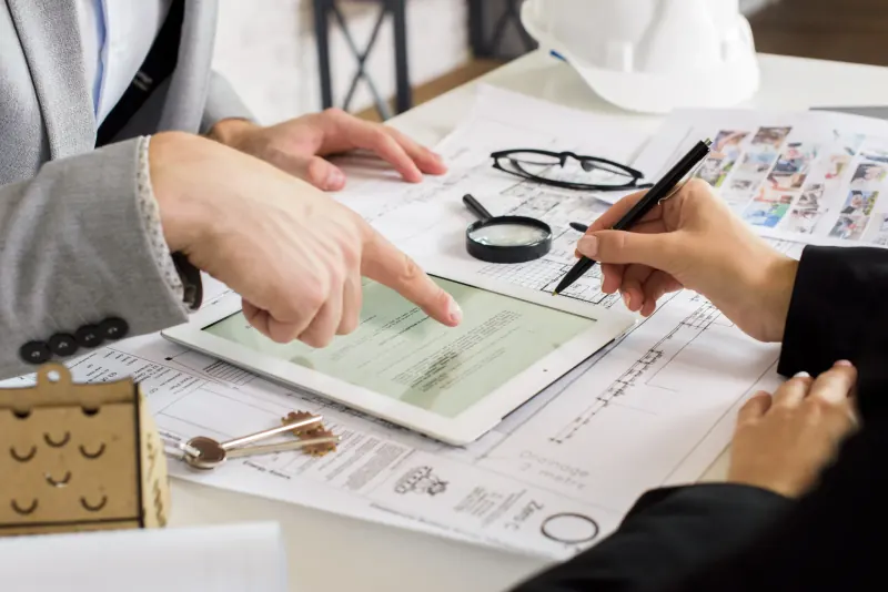 Hands pointing at a tablet over building plans, with a magnifying glass and house keys on the desk
