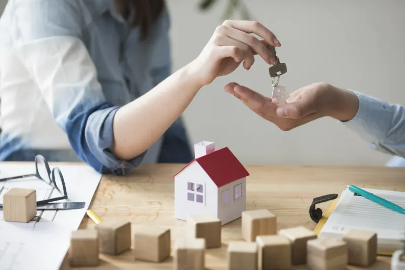 Hand passing a house key to another person over a table with a model house and wooden blocks