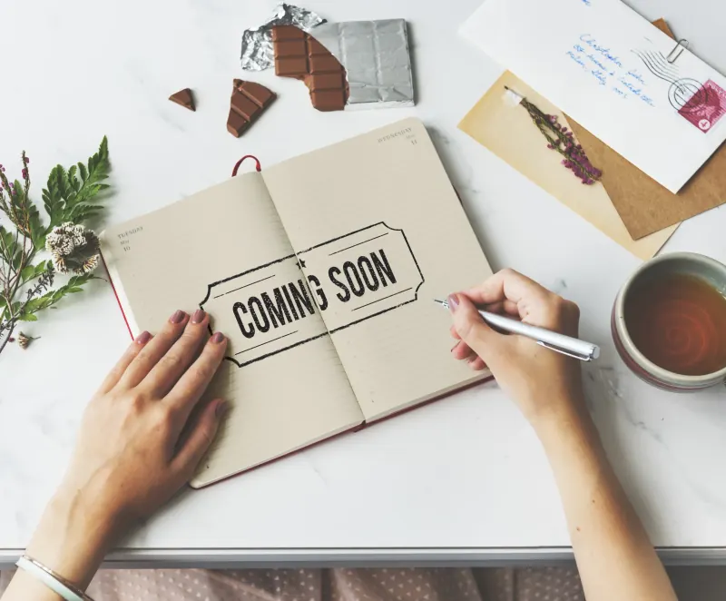 Hand writing “Coming Soon” in an open notebook on a marble desk beside a cup of tea