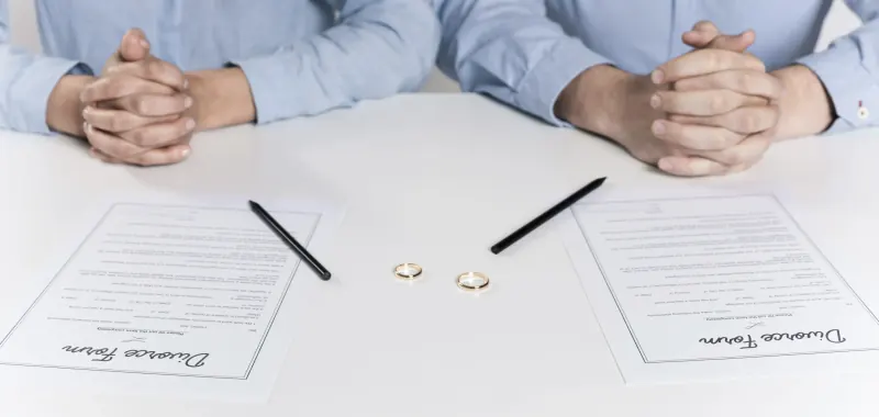 Couple seated at a table with two divorce forms, pens and their wedding rings in front of them