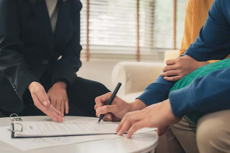 Couple signing paperwork on a clipboard while an agent guides them through the document