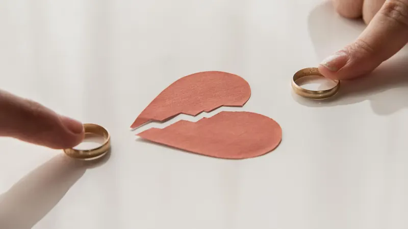 Two hands holding wedding rings on either side of a paper heart torn in two