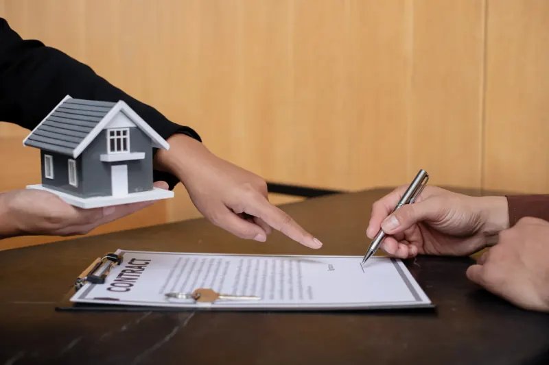Agent holding a model house and pointing to a real estate contract as a client signs it