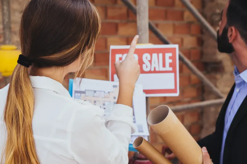 Woman pointing at a For Sale sign while reviewing building plans with a colleague
