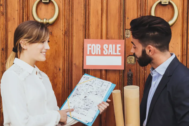 Two real estate agents reviewing a property plan at a front door with a For Sale sign